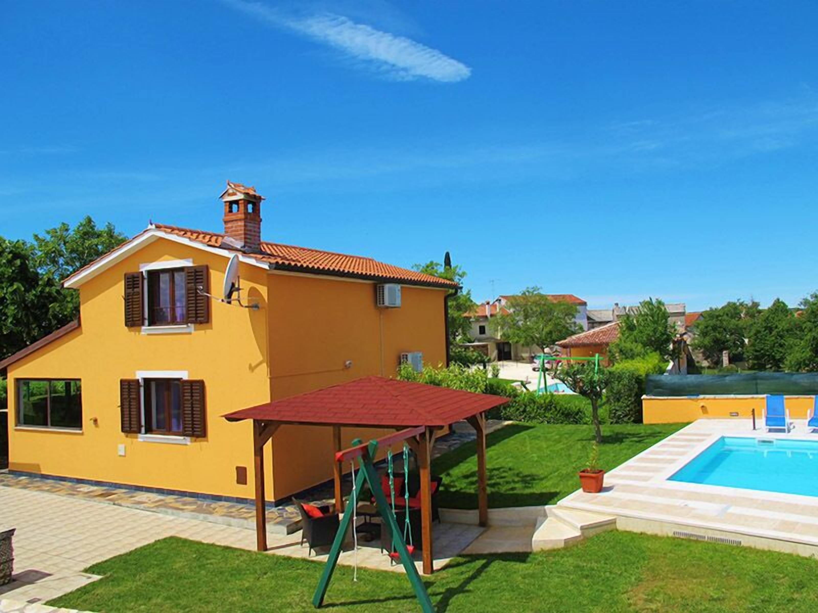 A yellow house with a red roof, a patio with a swing set, and a swimming pool.