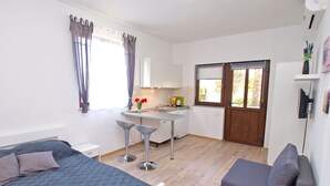 A bedroom with a bed, a small desk, and a chair. There is a window with curtains, a door, and a view of the outside.