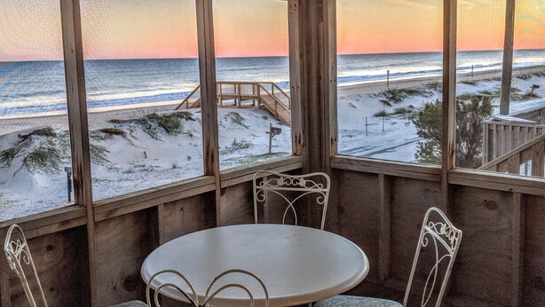 A beach view from a room with a round table and two chairs, a wooden deck, and a staircase leading to the beach.