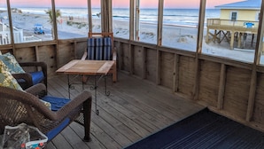 A covered deck with a view of the beach, a wooden table, wicker chairs, and a blue cushioned bench.