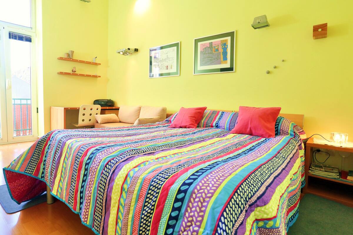 A bedroom with a colorful quilted bedspread, a wooden nightstand with a lamp, and a view of a balcony through a sliding door.