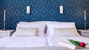 A neatly made bed with white linens, two bedside tables with candles, and a blue patterned wallpaper.