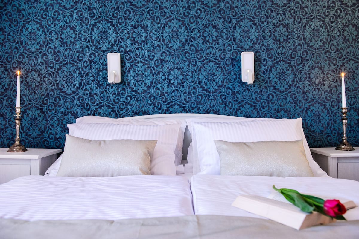 A neatly made bed with white linens, two bedside tables with candles, and a blue patterned wallpaper.