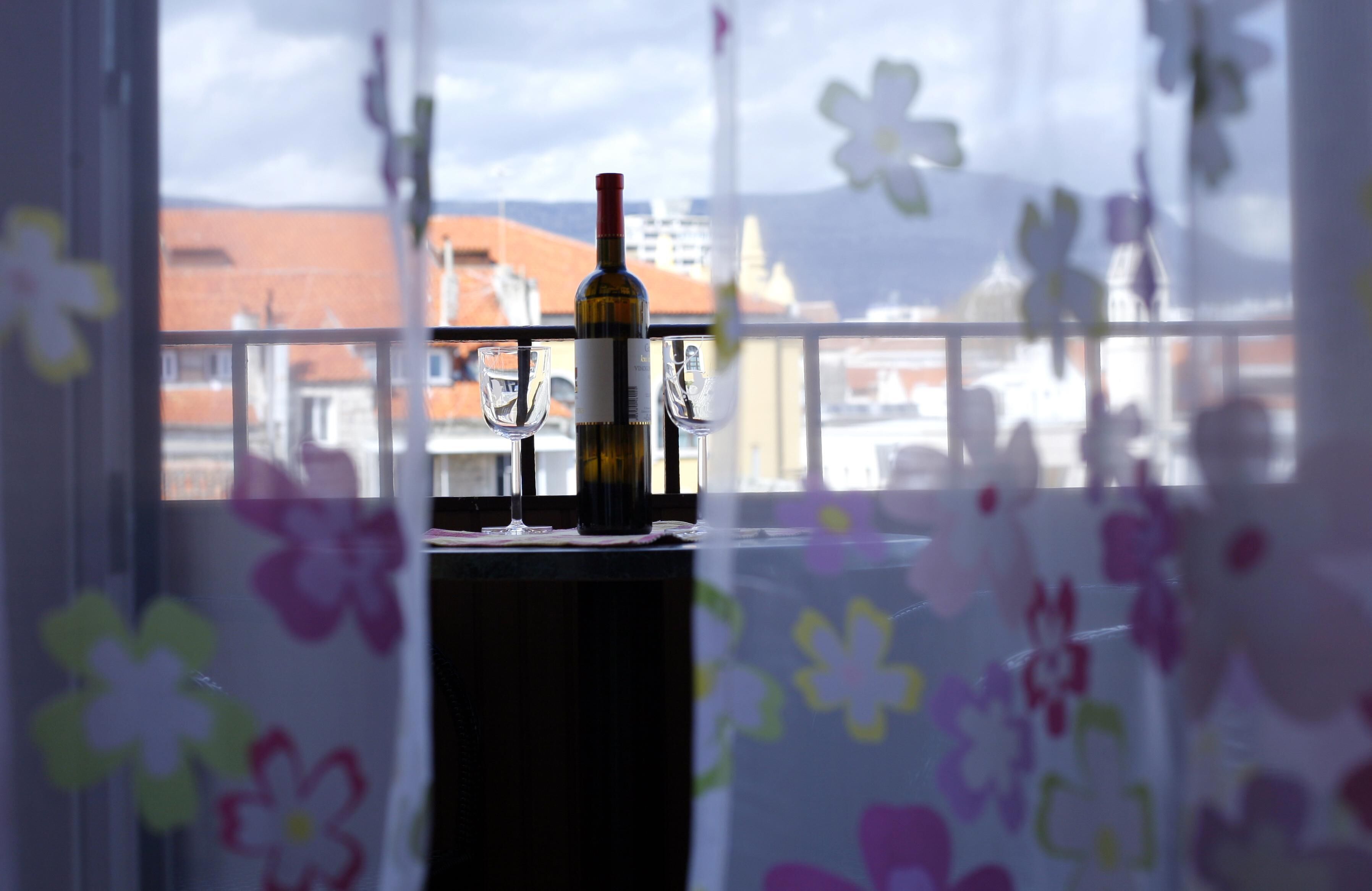 Un balcón con una botella y copas, vista de edificios y una ventana decorada con motivos florales.
