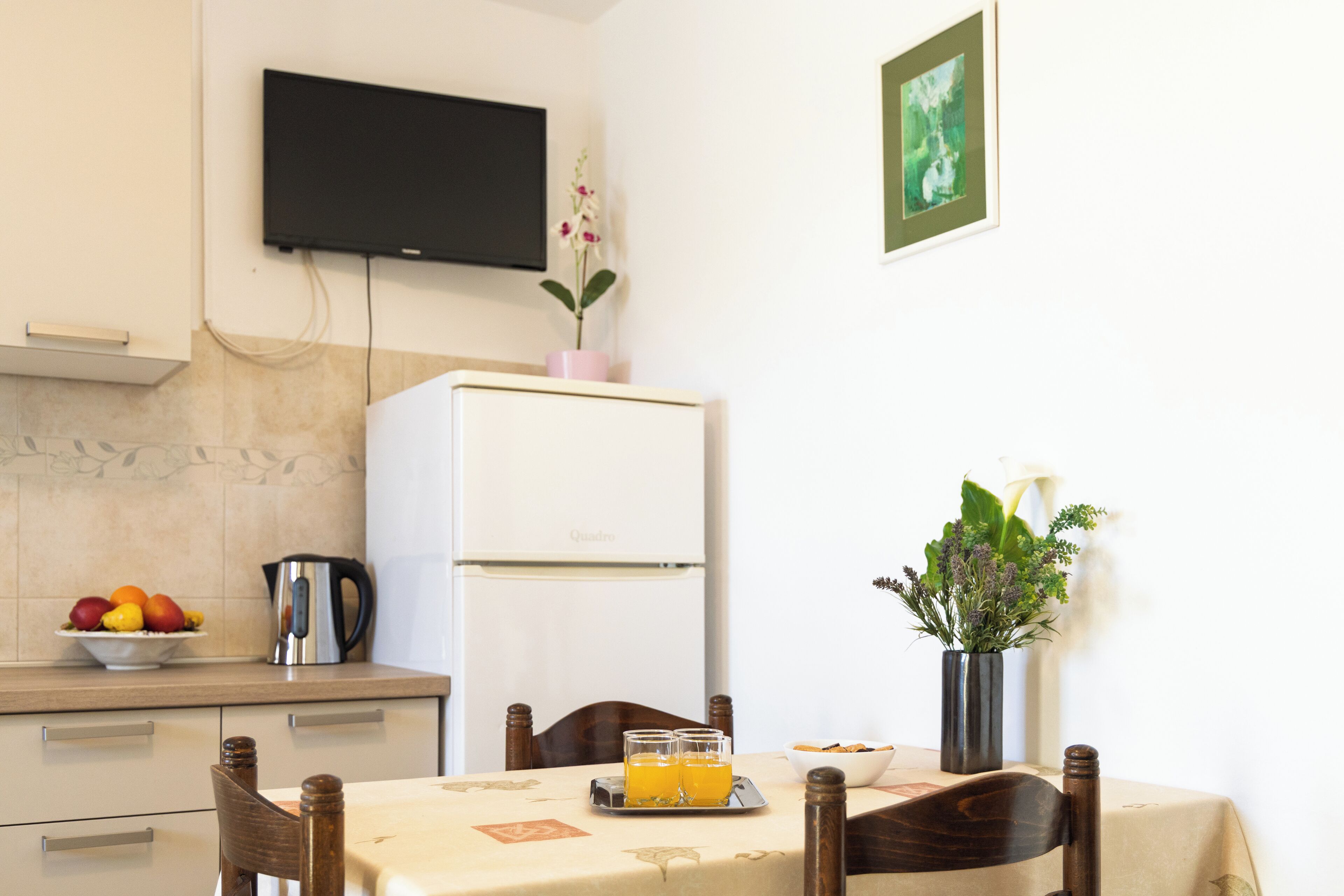 Una zona de menjador amb una taula preparada per a dues persones, un frigorífic blanc, un televisor fixat a la paret i un bol de fruita a la barra.