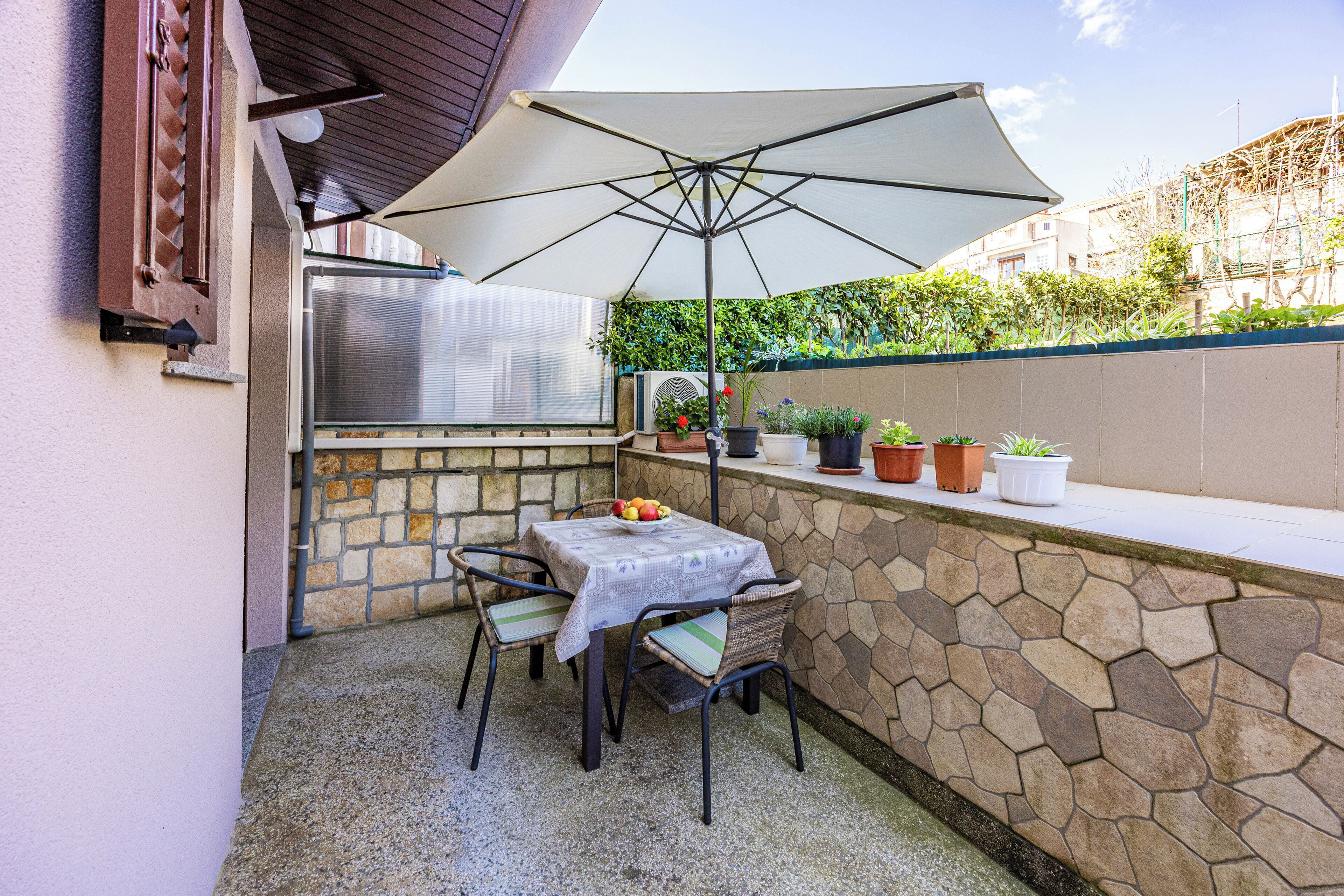 En udendørs terrasse med bord og stole, parasol, grill og krukker med planter.