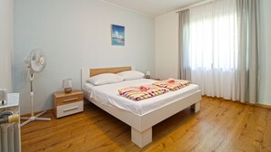 A bedroom with a wooden floor, a bed with white bedding, a bedside table with a lamp, a fan, and a window with curtains.