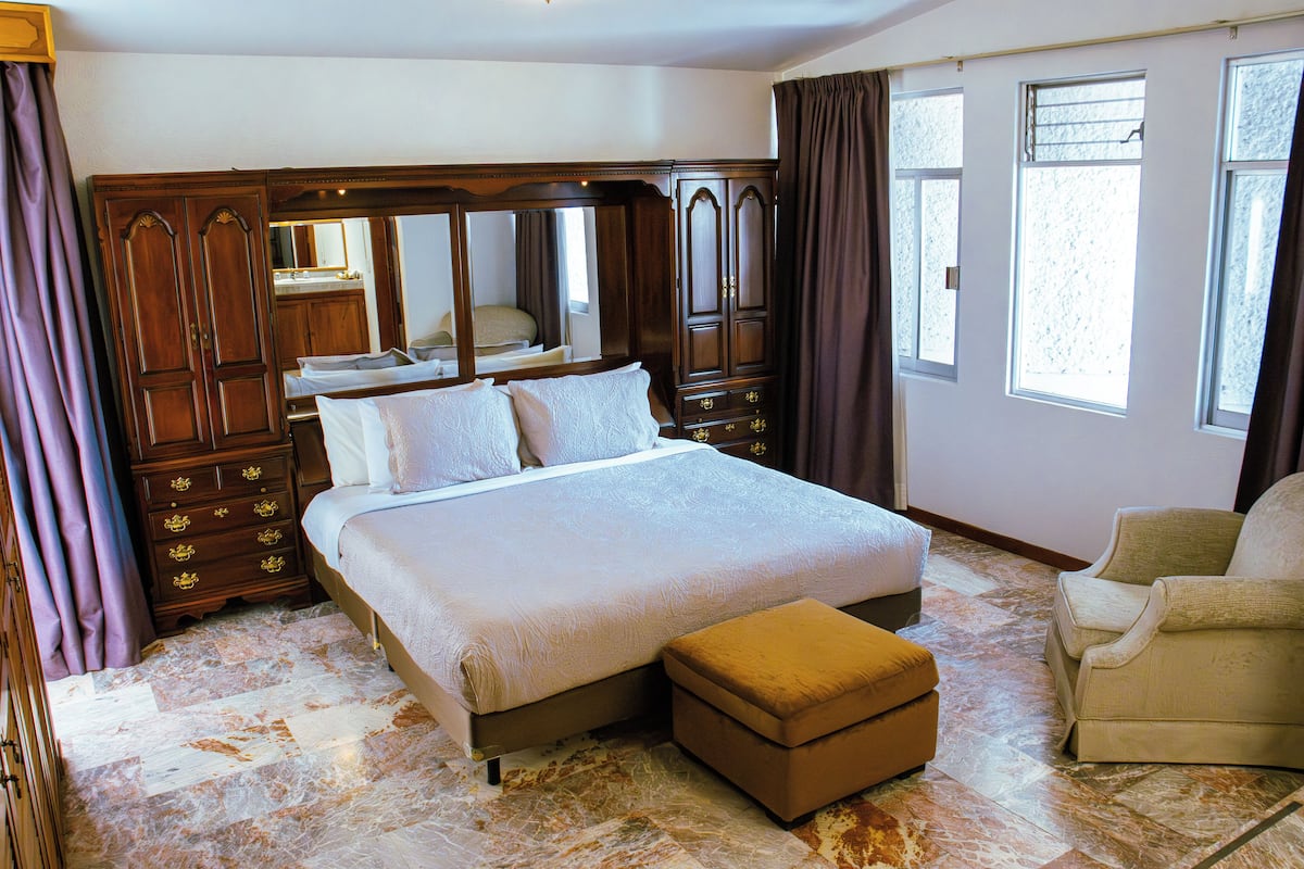 A spacious bedroom with a large bed, a wooden wardrobe, a sitting area with a chair, and a large window providing natural light.
