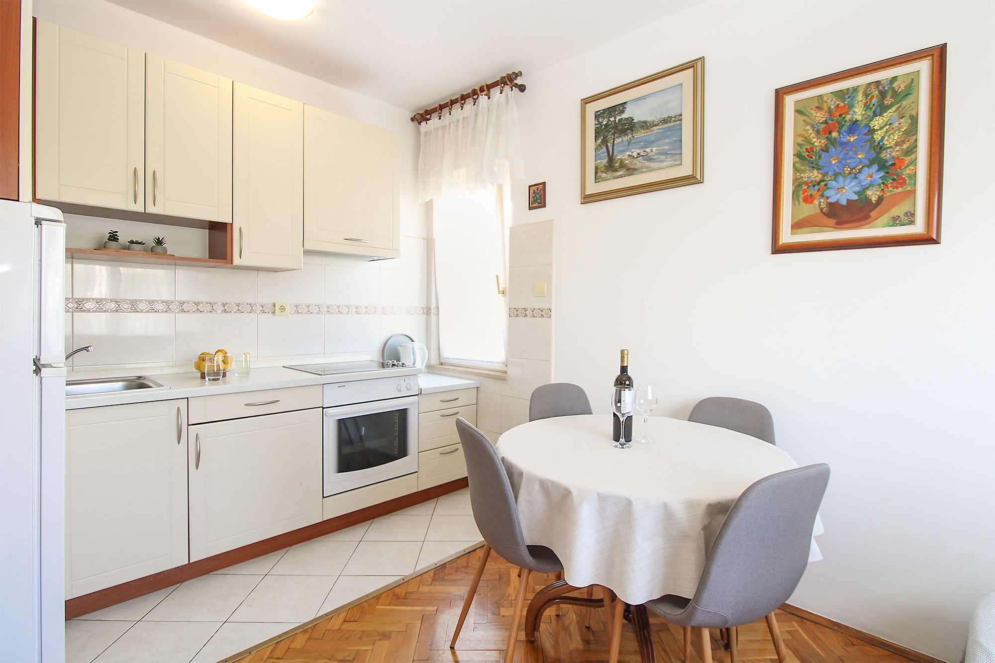 Une cuisine avec des meubles blancs, un évier, une cuisinière et une table à manger avec des chaises. Deux tableaux encadrés sont accrochés au mur.