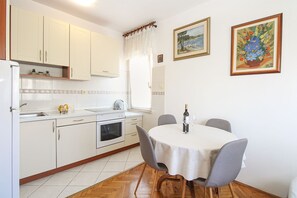Een keuken met witte kasten, een spoelbak, een fornuis en een eettafel met stoelen. Twee ingelijste schilderijen hangen aan de muur.