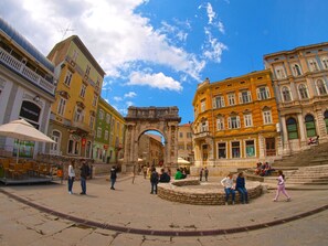 Une place historique, ornée d’une arche, entourée de bâtiments aux couleurs vives, et une fontaine en pierre au centre.