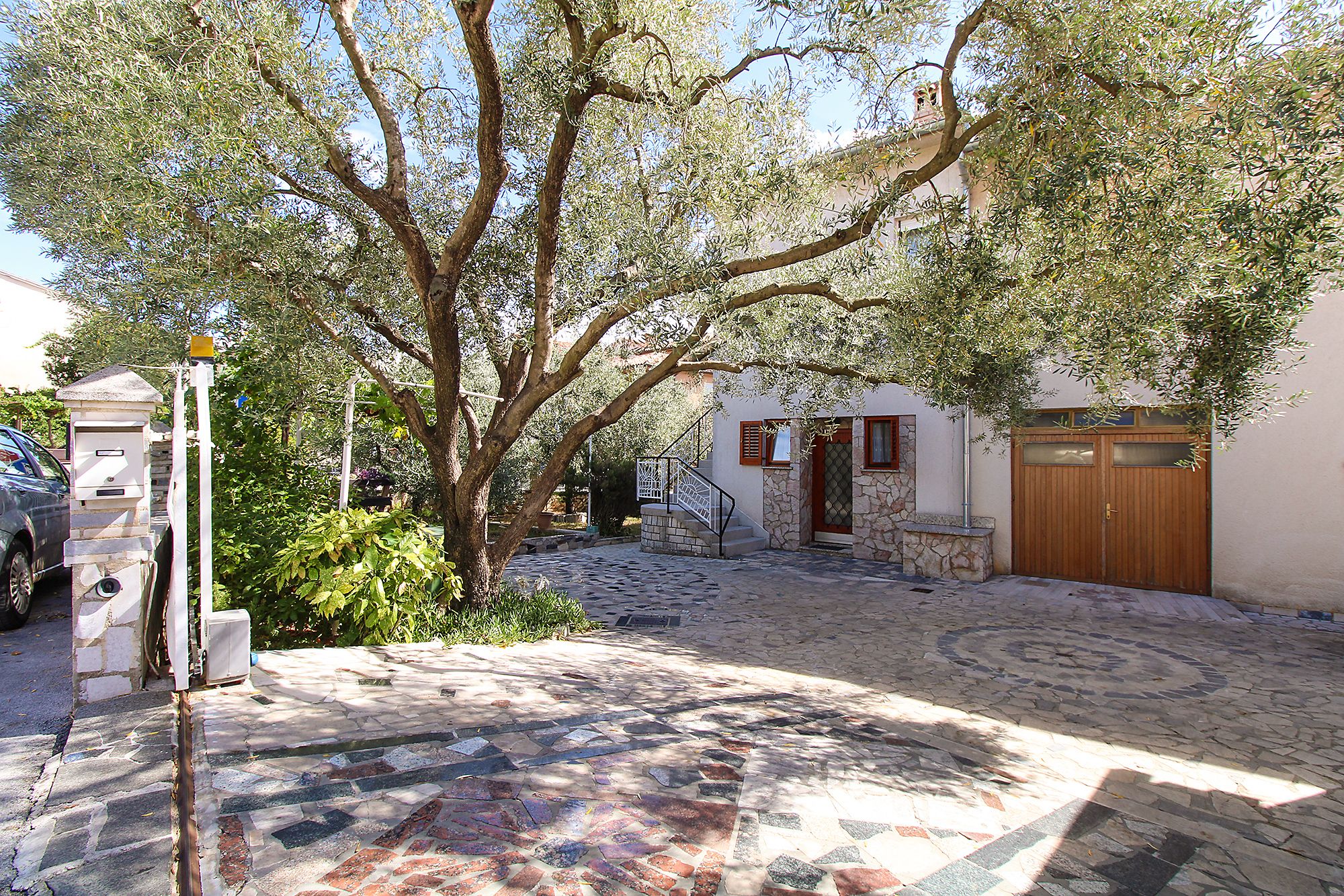 A courtyard with a stone-paved area, a tree, a building with a wooden door, and a vehicle parked outside.
