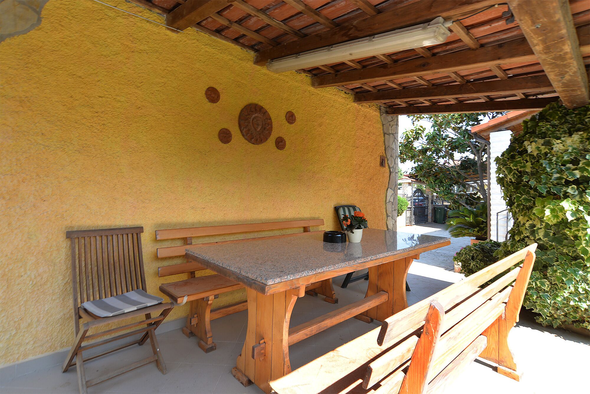 An outdoor patio with wooden benches and a table, a yellow wall with decorative plates, and a view of greenery.