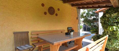An outdoor patio with wooden benches and a table, a yellow wall with decorative plates, and a view of greenery.