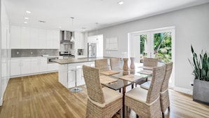 A modern kitchen with white cabinets, a central island, and a dining area with wicker chairs and a glass table. - Modern Oasis in Tropical Serene Victoria Park/Private Pool/Near Beach Las Olas (Fort Lauderdale)