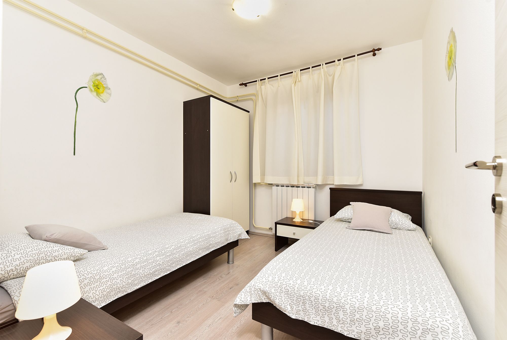 A bedroom with two single beds, a bedside table with a lamp, a wardrobe, and a window with curtains.
