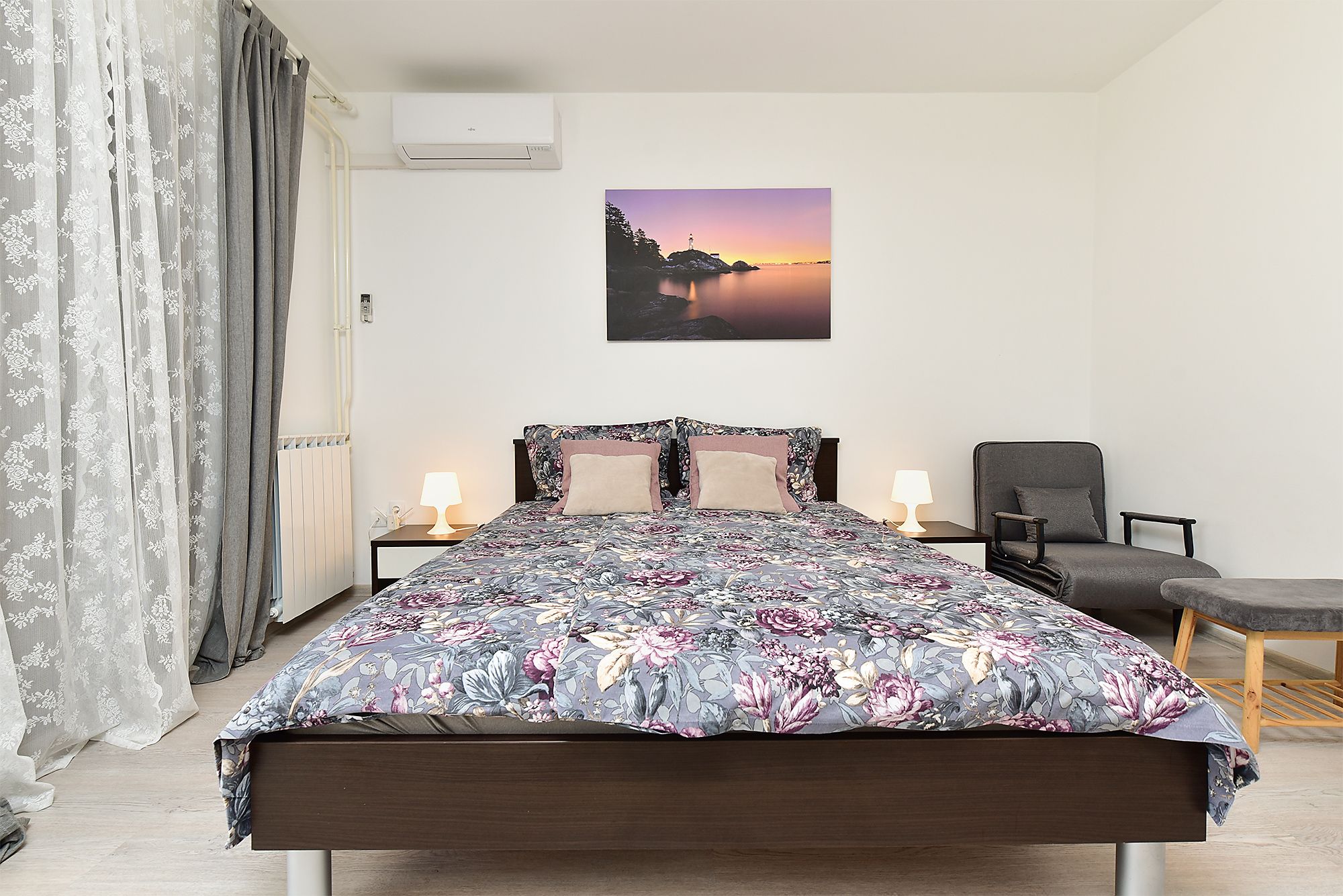Ein Schlafzimmer mit einem Bett, zwei Nachttischlampen, einem Sessel, einem Fenster mit Vorhängen und einer Wand-Klimaanlage.