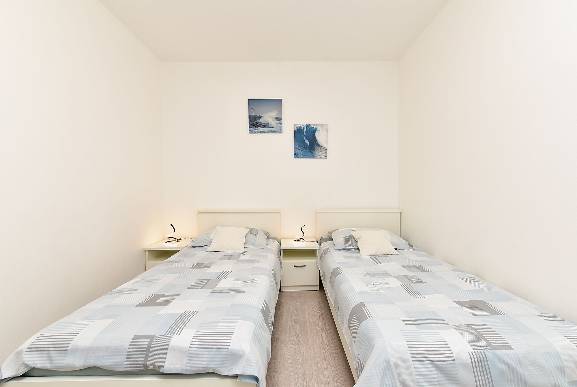 A bedroom with two single beds, a nightstand with a lamp, and two wall-mounted pictures.