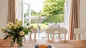 A dining area with a wooden table, a vase of flowers, a plate with a cake and blueberries, and a newspaper. - Fourways - Four Bedroom Cottage, Sleeps 8 (Townshend)