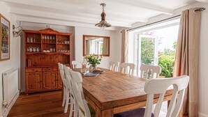 A dining area with a wooden table, white chairs, a wooden cabinet with glassware, and a view of greenery outside. - Fourways - Four Bedroom Cottage, Sleeps 8 (Townshend)