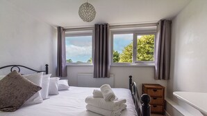 A neatly made bed with white linens and a rolled towel, a wooden nightstand, a radiator, and a window with curtains. - Fourways - Four Bedroom Cottage, Sleeps 8 (Townshend)
