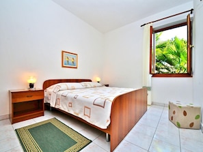 A bedroom with a bed, bedside lamps, a nightstand, a green patterned rug, a window with a view of greenery, and a framed picture on the wall. - Apartment 653/1563 (Istria - Rabac), 500m from the beach (Rabac)