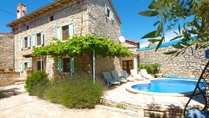 Ein Gebäude aus Stein mit Pool, Sitzgelegenheiten im Freien und einer Pergola, die mit grünen Weinreben bewachsen ist.