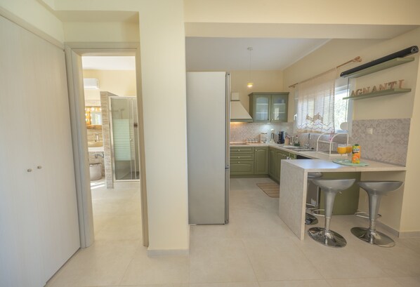 A modern kitchen with a refrigerator, bar stools, and a countertop with a fruit bowl and a bottle.