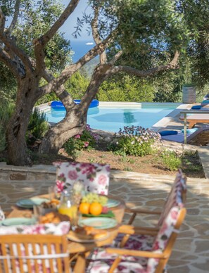 Un espace aménagé au bord de la piscine, avec une table dressée pour les repas, entouré de chaises longues et d’un grand arbre.
