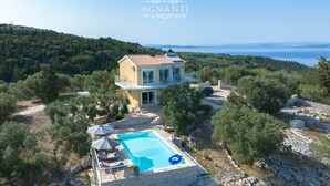 Une villa à deux étages avec piscine, entourée d’arbres et offrant une vue sur la mer.