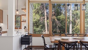 Eine moderne Küche mit weißen Schränken, einem Holztisch und einem großen Fenster mit Blick auf einen Wald.