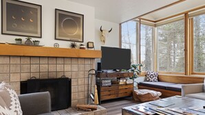 Ein gemütliches Wohnzimmer mit Kamin, Fernseher und großem Fenster mit Blick auf Bäume.
