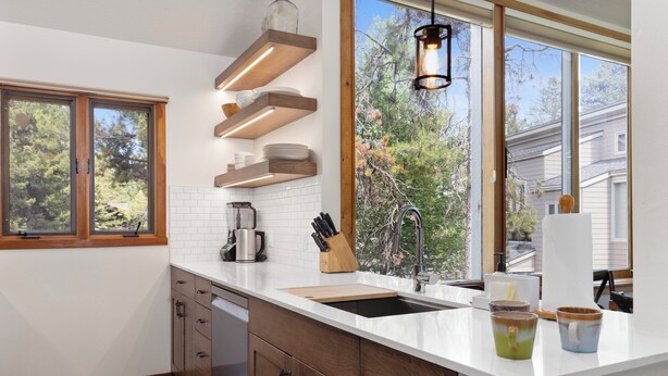 Eine moderne Küche mit Holzausstattung, einer weißen Arbeitsplatte, einem eingebauten Ofen und einem großen Fenster mit Blick auf Bäume und Häuser.