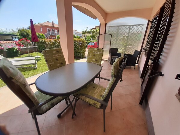 Un patio couvert avec une table et des chaises, donnant sur un jardin, et avec un bâtiment visible au loin.
