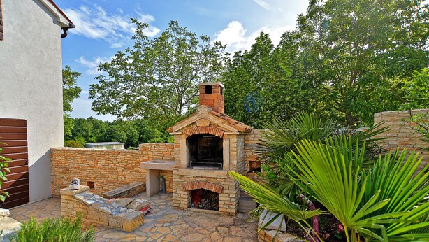 Une terrasse en pierre avec un four extérieur en briques, entourée de verdure et d’un bâtiment blanc.
