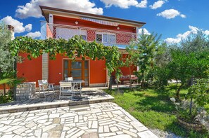 A two-story building with a red facade, a balcony with white railings, and a stone patio with outdoor furniture.