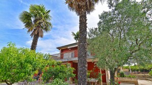 A building with a red facade, surrounded by various trees and plants, including a palm tree and a rose bush. - Apartment 684/1615 (Istria - Stinjan), 400m from the beach (Stinjan)
