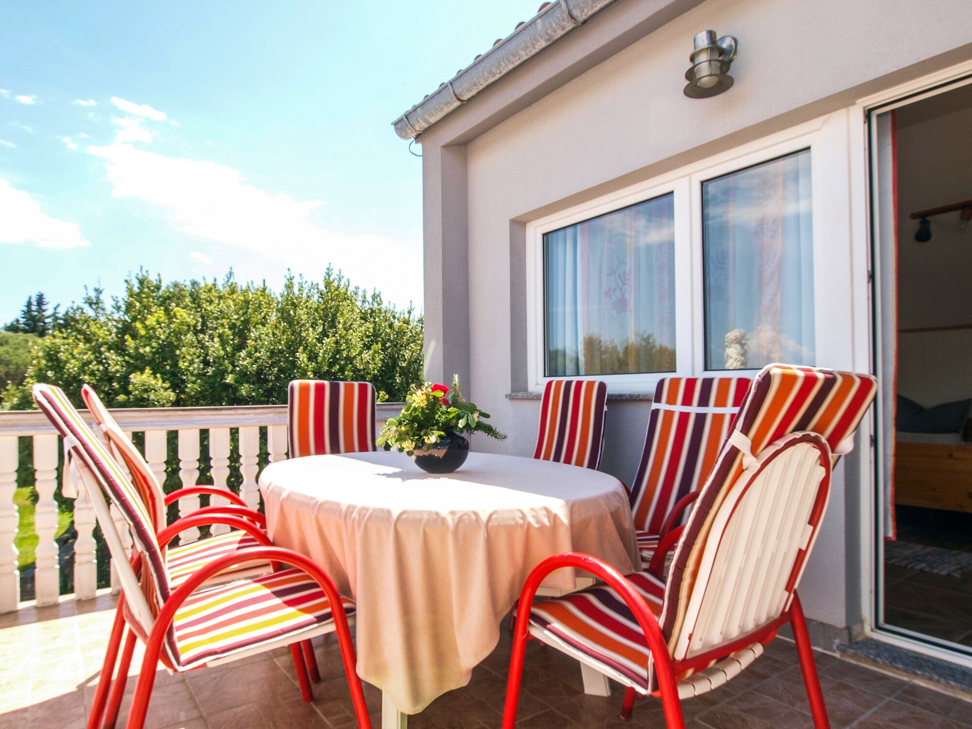 A round table with striped chairs on a balcony, a potted plant, and a view of greenery and a building in the background.