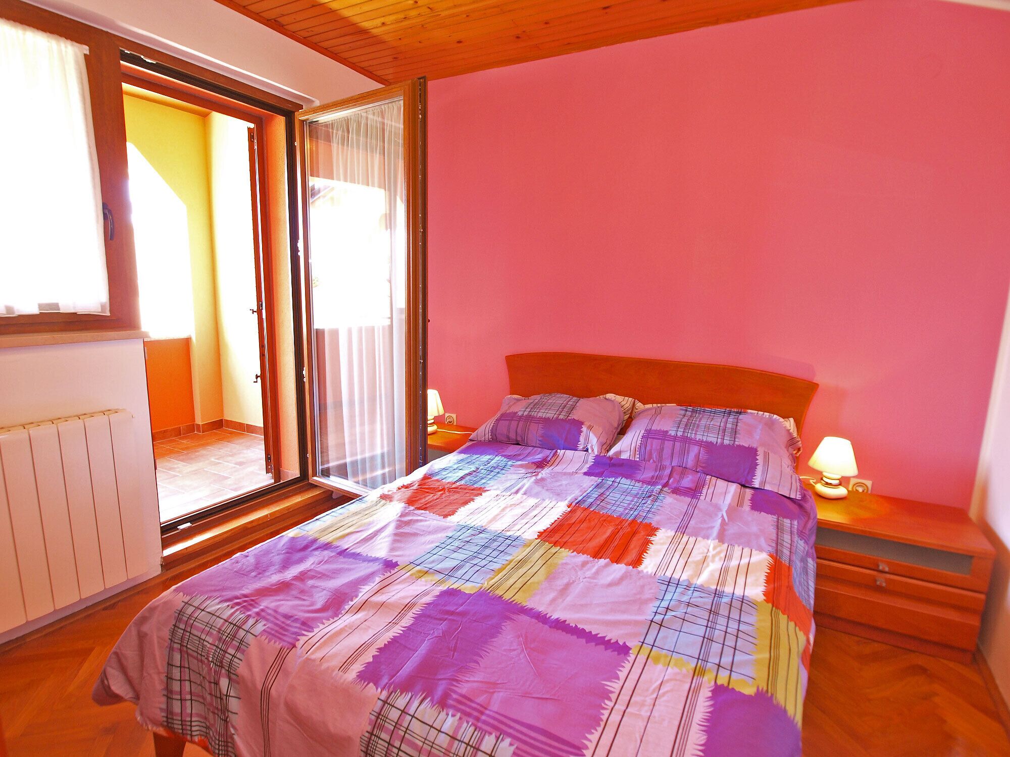 A bedroom with a large bed, a wooden headboard, a nightstand with a lamp, and a sliding glass door leading to a balcony.