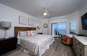 Ein Schlafzimmer mit einem Bett, einem Deckenventilator, einem Nachttisch, einer Kommode und Meerblick durch eine Schiebetür.