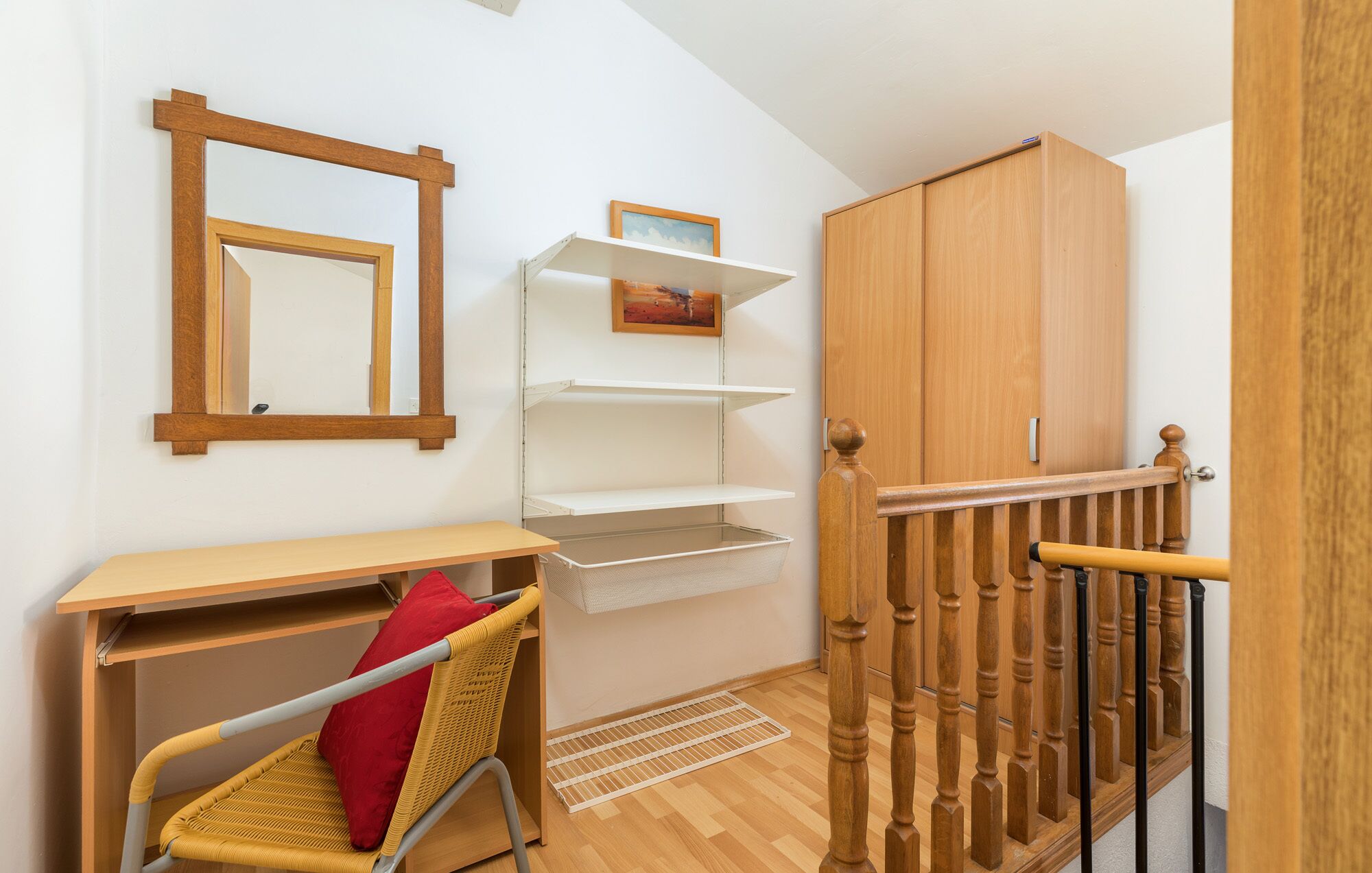 A room with a wooden desk, a wicker chair, a mirror, a wooden wardrobe, and a staircase with a wooden railing.