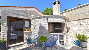 A stone-built outdoor kitchen area with a chimney, two blue chairs, and potted plants.