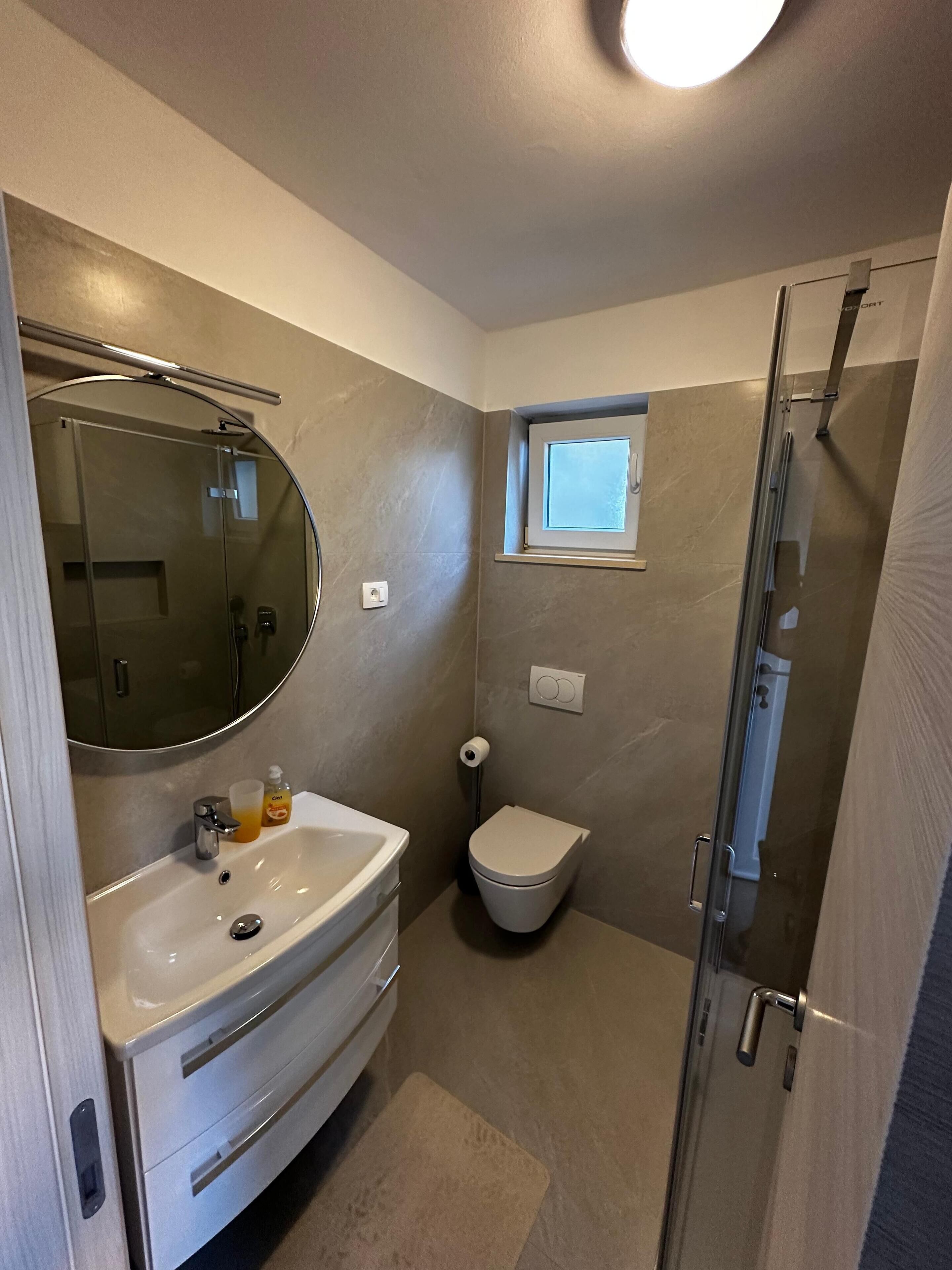 Une petite salle de bain avec un lavabo blanc, des toilettes, une cabine de douche en verre et une petite fenêtre située au-dessus des toilettes.