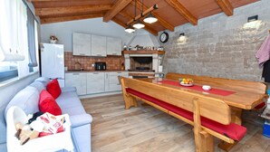 A cozy kitchen with wooden beams, a dining table with benches, a sofa with red cushions, and a stone wall.