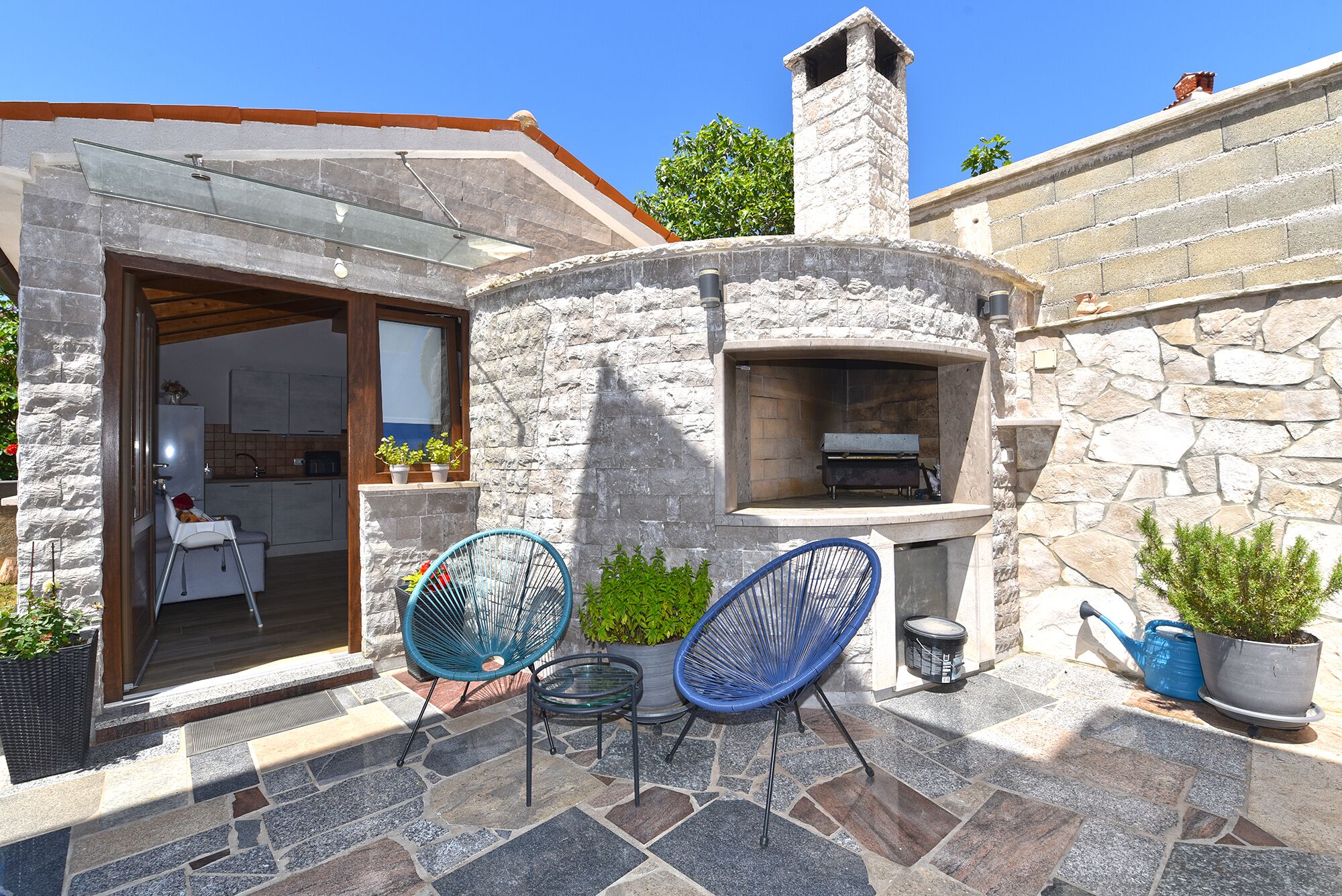 A stone-built outdoor kitchen area with a chimney, two blue chairs, and potted plants.