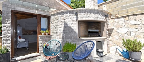 A stone-built outdoor kitchen area with a chimney, two blue chairs, and potted plants.