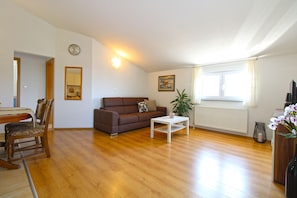 Una sala de estar con un sofá marrón, una mesa de centro blanca, una mesa de comedor con sillas, un reloj en la pared y una ventana con cortinas.