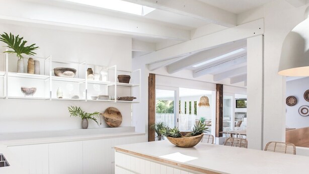 Una cocina moderna con armarios blancos, encimera de madera y una isla central con un cuenco de piñas. Estantes sobre la encimera exhiben diversos utensilios de cocina y objetos decorativos.