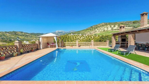 A rectangular swimming pool with a tiled deck, surrounded by a brick wall and a pergola with white curtains. There are lounge chairs and a barbecue grill on the right side.
