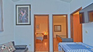 Een kleine kamer met een bed, een slaapbank en een eettafel. Aan de muur hangt een kunstwerk en er is een raam met zonwering.